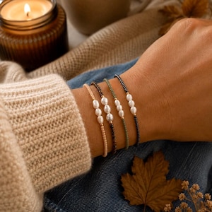 Könnte beinhalten: Ein Set aus vier Perlenarmbändern, die jeweils drei kleine weiße Süßwasserperlen in der Mitte eines Strangs aus winzigen Perlen aufweisen. Die Armbänder sind in den Farben Hellbeige, Dunkelbraun, Olivgrün und Schwarz mit goldenen Akzenten gehalten.