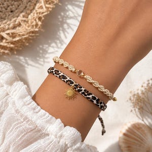Könnte beinhalten: Ein Set aus zwei Armbändern. Eines besteht aus einer beigen Makramee-Kordel mit einer kleinen, klaren Kristallperle. Das zweite ist ein flaches Band mit braun-schwarzem Leopardenmuster, akzentuiert durch einen goldenen Sonnenanhänger.