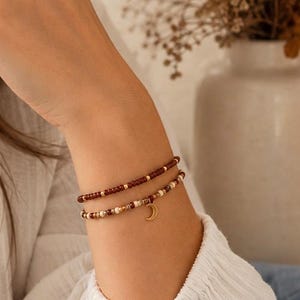 Könnte beinhalten: Zwei Perlenarmbänder, die zusammen getragen werden. Das obere Armband besteht aus kleinen dunkelroten Perlen mit goldenen Zwischenelementen. Das untere Armband kombiniert cremefarbene, braune und goldene Perlen und hat einen kleinen goldenen Mondsichelanhänger.