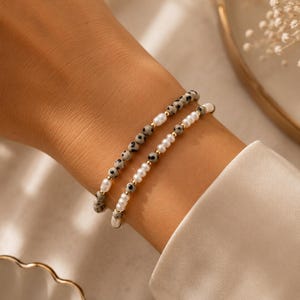 Könnte beinhalten: Zwei Perlenarmbänder bestehend aus einer Kombination kleiner weißer Perlen, gesprenkelter Dalmatiner-Jaspis-Perlen und winzigen goldfarbenen Metallzwischenstücken. Die Armbänder sind auf einem flexiblen Band aufgezogen und geschichtet.