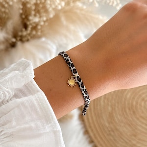 Könnte beinhalten: Ein schmales Armband mit einem weißen Stoffband und einem schwarzen sowie braunen Leopardenmuster. Das Armband ist mit einem kleinen, runden, goldfarbenen Metallanhänger in Form einer Sonne mit strahlenden Strahlen am Stoffband verziert.