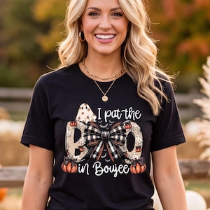 May include: A black short-sleeved t-shirt featuring the white text I put the BOO in Boujee with a central black and white plaid bow graphic, small orange pumpkin illustrations, and a ghost icon above the word BOO, paired with dark blue denim jeans.