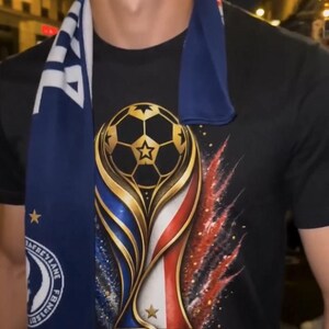 May include: Black t-shirt featuring a gold, blue, and red trophy design with a soccer ball and the text "FRANCE 2026". A blue scarf with white fringe and text is draped around the neck.