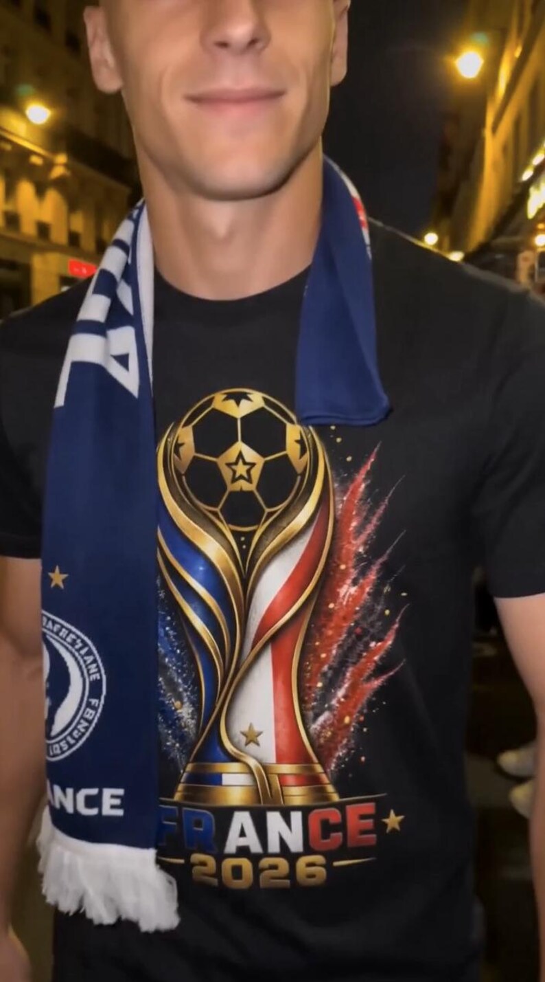 May include: Black t-shirt featuring a gold, blue, and red trophy design with a soccer ball and the text "FRANCE 2026". A blue scarf with white fringe and text is draped around the neck.