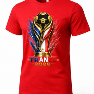 May include: Red t-shirt featuring a gold and black soccer ball trophy design with the text "FRANCE 2026". The design incorporates the colors of the French flag with blue, white, and red accents.
