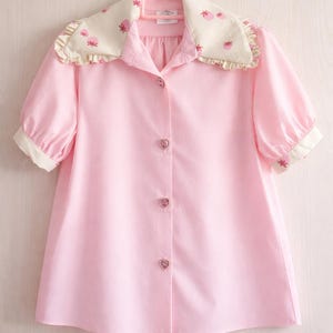 Peut inclure: Chemise rose clair à manches courtes bouffantes. Le col et les poignets sont crème avec un motif floral. Des boutons en forme de cœur ornent le devant. La chemise est suspendue à un cintre rose.