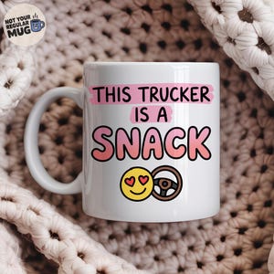 May include: A white ceramic mug featuring the black text THIS TRUCKER IS A SNACK with pink brushstroke accents. Below the text is a yellow smiley face emoji with heart eyes next to a brown steering wheel icon. A logo is in the top left corner.