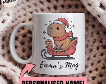 Può includere: Tazza in ceramica bianca con un capibara dei cartoni animati con cappello e sciarpa di Babbo Natale, su una slitta con luci. La tazza ha il testo "Emma's Mug" e "NOT YOUR REGULAR MUG". Un regalo personalizzato.