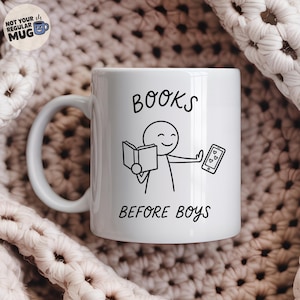 Op de afbeelding: Een witte keramische mok met een zwarte lijntekening van een stokfiguur die een open boek vasthoudt en een gebaar maakt naar een smartphone met hartjes. De tekst BOOKS BEFORE BOYS is in het zwart op de mok gedrukt.