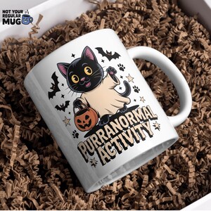 May include: White ceramic mug with a Halloween-themed design. The mug features a black cat dressed as a ghost, holding a jack-o'-lantern. The text "PURRANORMAL ACTIVITY" is printed below the cat, with bats and paw prints.