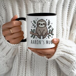 K&ouml;nnte beinhalten: Wei&szlig;er Keramikbecher mit schwarzem Innenraum und Henkel. Der Becher zeigt ein Cartoon-Faultier auf einem Ast, umgeben von Bl&auml;ttern und Blumen. Der Text "AARON'S MUG" ist darunter gedruckt.