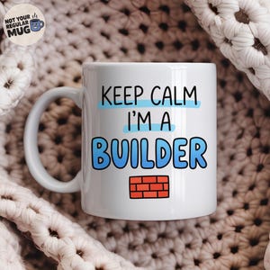 May include: A white ceramic mug featuring the black text "KEEP CALM I'M A BUILDER" with light blue horizontal accents behind the words and a small red brick wall graphic at the bottom. The mug has a curved handle and a smooth, glossy finish.