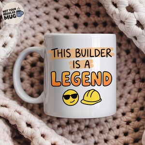 Könnte beinhalten: Ein weißer Keramikbecher mit dem schwarzen Text "THIS BUILDER IS A LEGEND" und orangen Akzenten. Unter dem Text befinden sich ein gelbes Smiley-Gesicht mit schwarzer Sonnenbrille und ein gelbes Bauhelm-Symbol. Ein Logo befindet sich oben links.