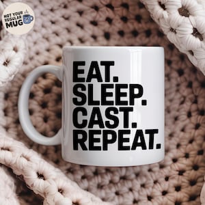 Puede incluir: Taza de cerámica blanca con el texto "EAT. SLEEP. CAST. REPEAT." en letras negras en negrita. La taza tiene un asa curva y un pequeño logotipo que dice "NOT YOUR REGULAR MUG". La taza está sobre un fondo texturizado blanquecino.