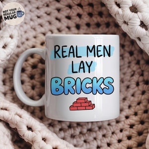 May include: A white ceramic mug featuring the black text "REAL MEN LAY BRICKS" with light blue brush stroke accents behind the words. Below the text is a small graphic of a red brick wall. The mug has a curved handle on the left side.