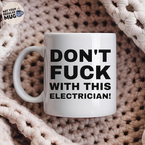 以下が含まれることがあります： 「DON'T FUCK WITH THIS ELECTRICIAN!」という黒い太字のテキストがサンセリフ体でプリントされた白いセラミック製マグカップ。左側に標準的な持ち手があり、滑らかで光沢のある仕上げで、メッセージがはっきりと読み取れるデザインになっています。