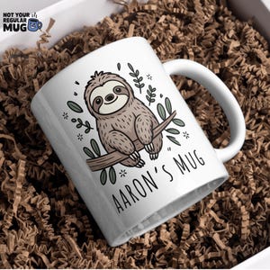 K&ouml;nnte beinhalten: Wei&szlig;e Keramik-Tasse mit einem Cartoon-Faultier auf einem Ast, umgeben von Bl&auml;ttern und Sternen. Der Text "AARON'S MUG" ist darunter gedruckt. Die Tasse wird in einer Box mit braunem Papierf&uuml;llmaterial pr&auml;sentiert.