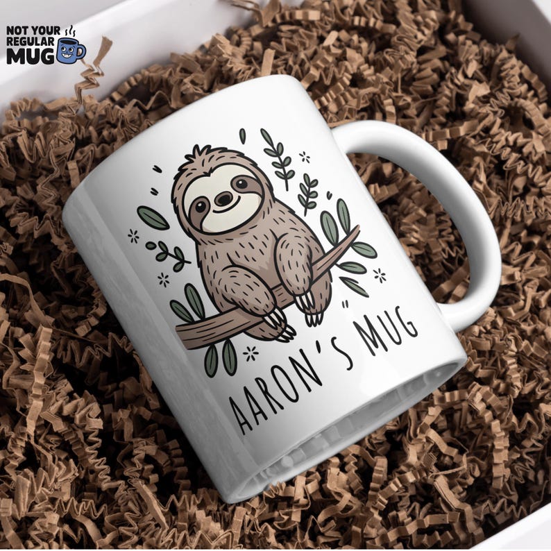 K&ouml;nnte beinhalten: Wei&szlig;e Keramik-Tasse mit einem Cartoon-Faultier auf einem Ast, umgeben von Bl&auml;ttern und Sternen. Der Text "AARON'S MUG" ist darunter gedruckt. Die Tasse wird in einer Box mit braunem Papierf&uuml;llmaterial pr&auml;sentiert.