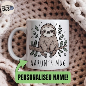 Könnte beinhalten: Weiße Keramiktasse mit einem Cartoon-Faultier auf einem Ast, umgeben von grünen Blättern und Sternen. Die Tasse zeigt "Aaron's Mug" und "Personalised Name!". Außerdem ist der Text "NOT YOUR REGULAR MUG" aufgedruckt.