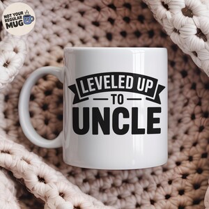 以下が含まれることがあります： 黒い文字で「LEVELED UP TO UNCLE」と書かれた白いセラミックマグ。バナーデザインです。マグには湾曲したハンドルがあり、テクスチャ加工されたオフホワイトの背景に置かれています。「NOT YOUR REGULAR MUG」と書かれた小さなロゴが左上にあります。