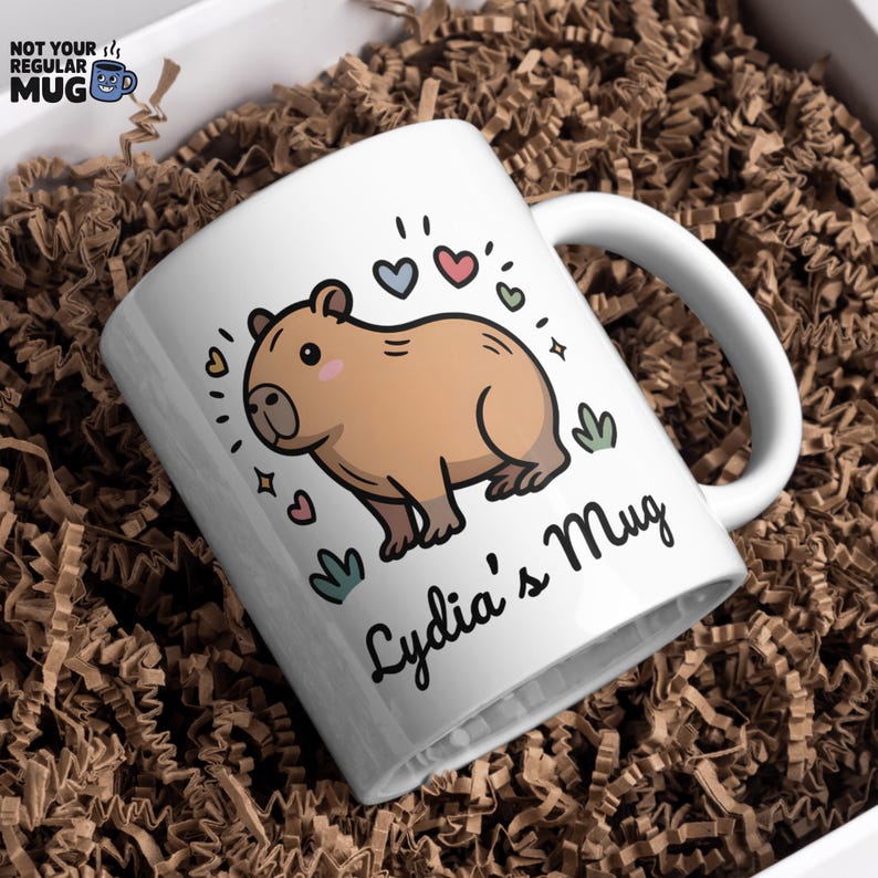 May include: A white ceramic mug featuring a cartoon illustration of a tan capybara surrounded by small colorful hearts and green leaf accents. The text Lydia's Mug is printed in black script font below the capybara illustration on the mug.