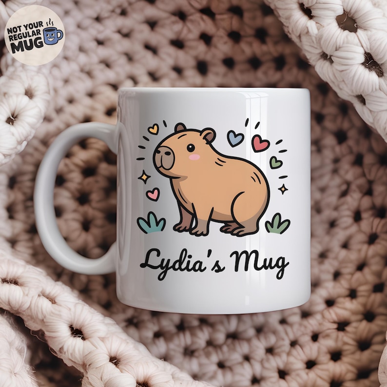 May include: A white ceramic mug featuring a cartoon illustration of a tan capybara surrounded by small colorful hearts and stars. Below the capybara, the text reads "Lydia's Mug" in a black script font. A small circular logo in the corner says "NOT YOUR REGULAR MUG".