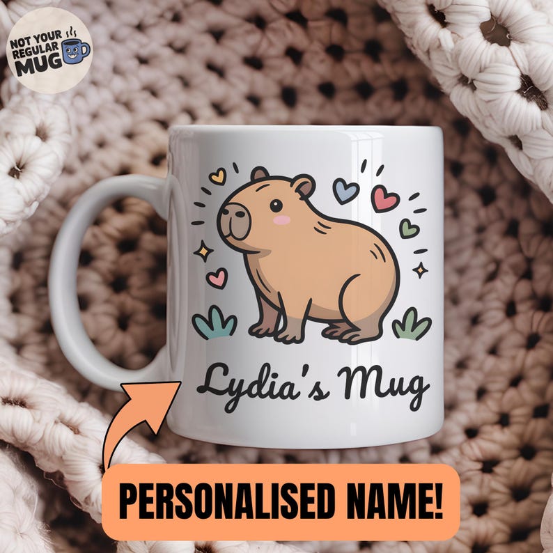 May include: A white ceramic mug featuring a cartoon illustration of a tan capybara surrounded by small colorful hearts and stars. The text "Lydia's Mug" is printed in black script below the capybara. An orange arrow points to the name.