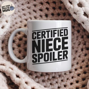 May include: White ceramic mug with the text "CERTIFIED NIECE SPOILER" in bold black letters. The mug has a curved handle and is set against a textured, off-white background. The mug is a standard size.