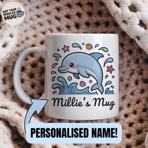 May include: White ceramic mug with a cartoon dolphin leaping from blue waves, decorated with starfish and shells. The mug is inscribed with "Millie's Mug" and the words "Personalised Name!" on a blue banner.