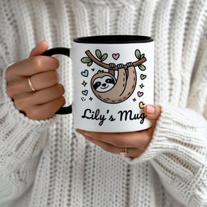 May include: White ceramic mug with a black interior and handle. The mug features a cartoon sloth hanging from a branch, surrounded by hearts and stars. The text "Lily's Mug" is written below the sloth in a script font.