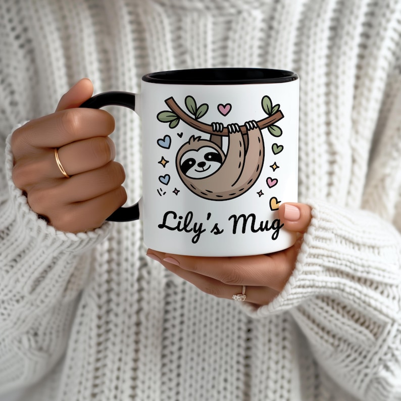 May include: White ceramic mug with a black interior and handle. The mug features a cartoon sloth hanging from a branch, surrounded by hearts and stars. The text "Lily's Mug" is written below the sloth in a script font.