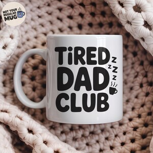 May include: White ceramic mug with the text "Tired Dad Club" in bold black letters. The design includes stylized Zzz's and a coffee cup icon. The mug has a curved handle and a round base.