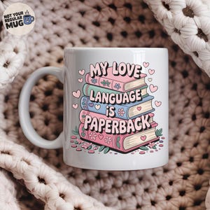 Op de afbeelding: Witte keramische mok met de tekst "MY LOVE LANGUAGE IS PAPERBACK" in roze, groen en blauw. Het ontwerp toont een stapel boeken met bloemen- en hartaccenten. De mok heeft een gebogen handvat en een klein logo.