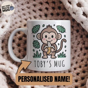 Peut inclure: Mug en céramique blanche avec un dessin de singe tenant une banane, entouré de feuilles et d'herbe vertes. Le mug porte l'inscription "TOBY'S MUG" et "NOM PERSONNALISÉ!". Il possède une anse incurvée.