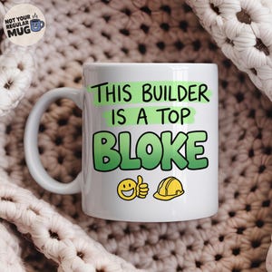 May include: A white ceramic mug featuring the black text THIS BUILDER IS A TOP BLOKE with green highlights. The design includes a yellow smiley face giving a thumbs up and a yellow construction hard hat icon below the text.