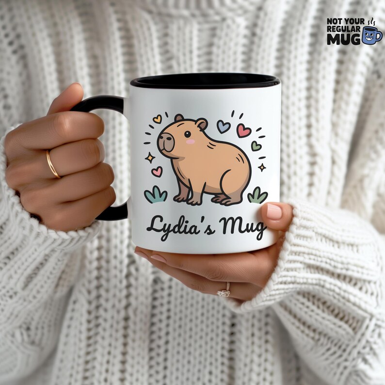 May include: A white ceramic mug with a black handle and interior rim. The front features a cartoon illustration of a tan capybara surrounded by small colorful hearts and stars. Below the capybara, the text reads Lydia's Mug in black script.