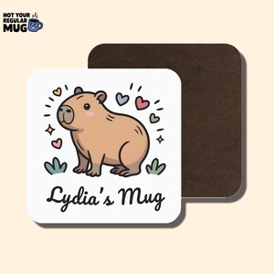 May include: A square coaster with a white top surface featuring a cartoon capybara, small colorful hearts, and the text "Lydia's Mug" in black script. The coaster has a dark brown underside and rounded corners, presenting a clean and simple design.