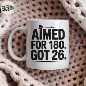 以下が含まれることがあります： 黒いダーツと「AIMED FOR 180. GOT 26」の文字が入った白いセラミックマグ。マグカップは湾曲したハンドルと丸い底を持っています。マグカップは、テクスチャ加工されたオフホワイトの表面にあります。