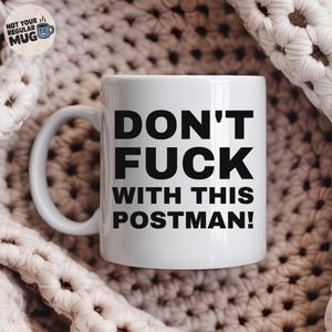 Può includere: Una tazza in ceramica bianca con un manico sul lato sinistro, caratterizzata da un testo nero in grassetto che recita "DON'T FUCK WITH THIS POSTMAN!" in lettere maiuscole. Un piccolo logo circolare nell'angolo in alto a sinistra mostra una tazza blu.