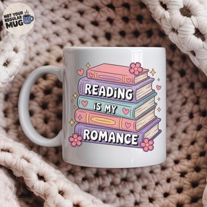 Może przedstawiać: Biały ceramiczny kubek z uchwytem, z kolorowym stosikiem książek z napisem "READING IS MY ROMANCE". Projekt zawiera serca, gwiazdy i kwiaty. Kubek ma napis "NOT YOUR REGULAR MUG" z boku.