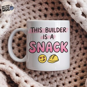 May include: A white ceramic mug featuring the black text THIS BUILDER IS A, followed by the pink text SNACK. Below the text are a yellow smiling emoji with heart eyes and a yellow construction hard hat. The mug has a curved handle on the left side.