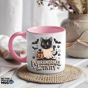 May include: White ceramic mug with a pink interior and handle. The mug features a Halloween-themed graphic of a black cat in a ghost costume holding a jack-o'-lantern. The text "PURRANORMAL ACTIVITY" is below the cat, with bats and stars.