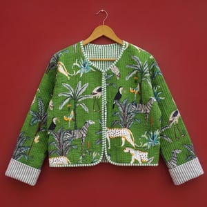 May include: A green quilted jacket featuring a vibrant jungle print with zebras, leopards, toucans, and palm trees. The garment has a V-neckline, long sleeves with grey and white striped cuffs, and matching striped trim along the front opening.