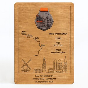 Op de afbeelding: Houten plaat met een zilveren medaille en een oranje lint. De medaille is gegraveerd met "DAM DVM 10KM" en stadsgezichten. De plaat bevat een racekaart, deelnemersgegevens en de tekst "DAM TOT DAMLOOP AMSTERDAM - ZAANDAM 21 september 2025".
