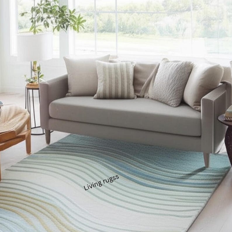 K&ouml;nnte beinhalten: Ein graues Sofa mit mehreren beigen Kissen steht &uuml;ber einem Teppich mit einem wellenf&ouml;rmigen Muster in Blau-, Gr&uuml;n- und Wei&szlig;t&ouml;nen. Ein Beistelltisch mit einer Lampe und ein Stuhl sind sichtbar. Der Teppich tr&auml;gt den Text "Living rugss".