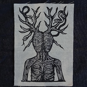 Può includere: Una toppa in bianco e nero raffigurante una figura a due facce con più occhi e rami simili a corna. I serpenti sono intrecciati nei rami. Anche il busto della figura è adornato da occhi. La toppa è rettangolare e ha uno sfondo grigio chiaro.