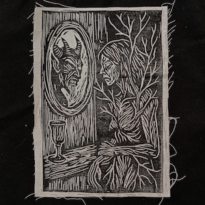 May include: Black and white linocut print on fabric. The artwork depicts a figure with tree-like features seated before a mirror reflecting a horned demon. A goblet sits on a table. The print has a raw, unfinished edge.