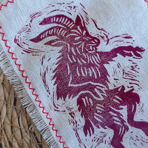 Könnte beinhalten: Ein quadratisches, cremefarbenes Leinentextil mit einem magentafarbenen Druck eines gehörnten Wesens. Die Kanten sind mit einem roten Zickzackstich und ausgefransten Kanten versehen. Das Bild ist eine Nahaufnahme, die die Textur des Stoffes und das kräftige Design hervorhebt.