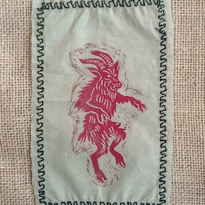 Puede incluir: Un parche de tela verde claro con una ilustración roja de una criatura parecida a una cabra. El parche tiene un borde cosido en zigzag verde oscuro. El fondo es una tela de arpillera beige.