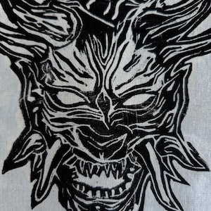 May include: A black and white illustration of a Japanese Oni mask. The mask has sharp horns, a wide grin with visible teeth, and detailed features. The artwork is set against a white background, creating a striking contrast.